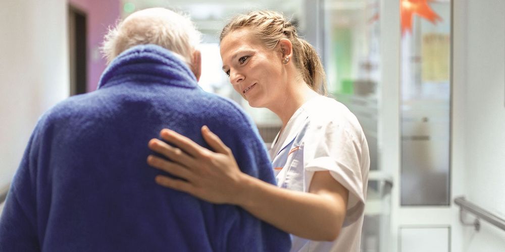 Eine Krankenpflegerin legt einem Senioren eine Hand auf den Rücken und begleitet diesen durch den Krankenhausflur.