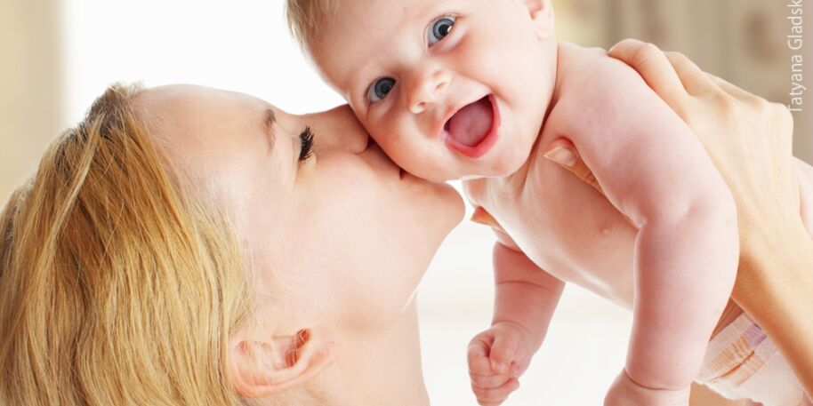 Eine Mutter, welche ihr Baby hoch vor ihr Gesicht hält und ihrem Baby einen Kuss auf die Wange drückt. Das Baby lacht. 