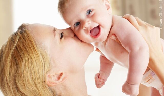 Eine Mutter, welche ihr Baby hoch vor ihr Gesicht hält und ihrem Baby einen Kuss auf die Wange drückt. Das Baby lacht.