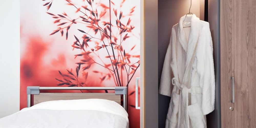 Bett in einem Zimmer des Krankenhaus Neuwerk. Auf der Tapete hinter dem Bett ist eine Pflanze. Neben dem Bett hängt ein Bademantel. 
