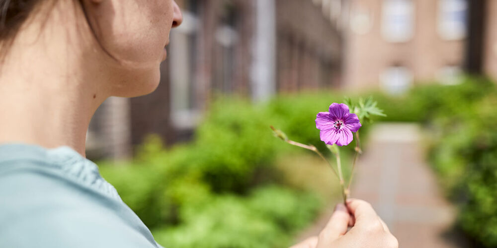 Eine Frau steht draußen vor einem Gebäude auf einem Weg und hält eine einzelne Blume in den Händen vor sich. 
