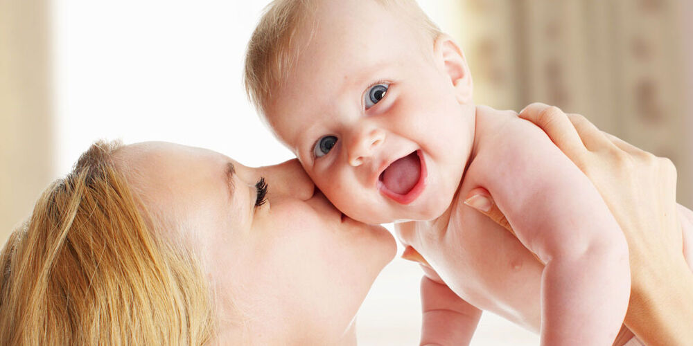 Eine Mutter küsst ihr lachendes Baby auf die Wange und hält es hoch. Das Baby lacht in die Kamera.