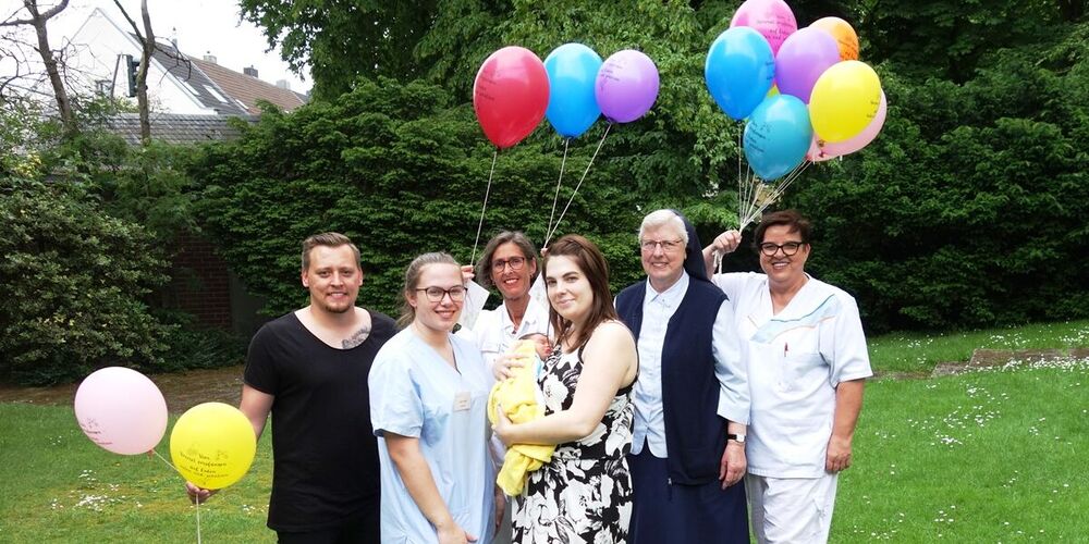 Garten in dem eine junge Familie mit ihrem frischgeborenem Baby steht. Neben dieser stehen auch noch drei Krankenpflegerinnen und eine Nonne in diesem Garten. Sie haben einige bunte Luftballons in der Hand.