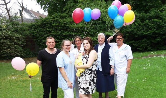 Garten in dem eine junge Familie mit ihrem frischgeborenem Baby steht. Neben dieser stehen auch noch drei Krankenpflegerinnen und eine Nonne in diesem Garten. Sie haben einige bunte Luftballons in der Hand.