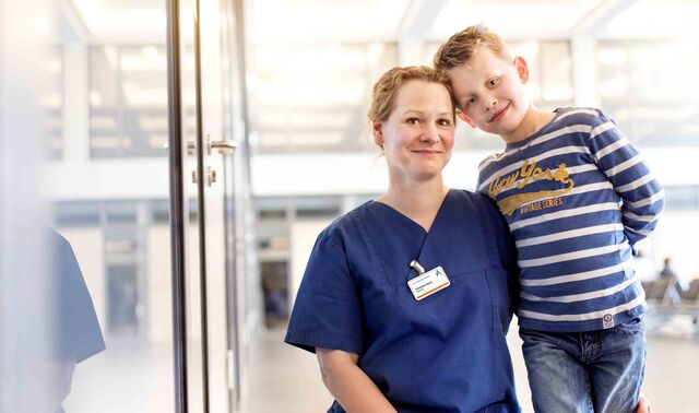 Eine Frau in Krankenhauskleidung kniet neben einem kleinen Jungen, welcher sich mit dem Kopf und Oberkörper an diese lehnt. Beide schauen lächelnd in die Kamera.