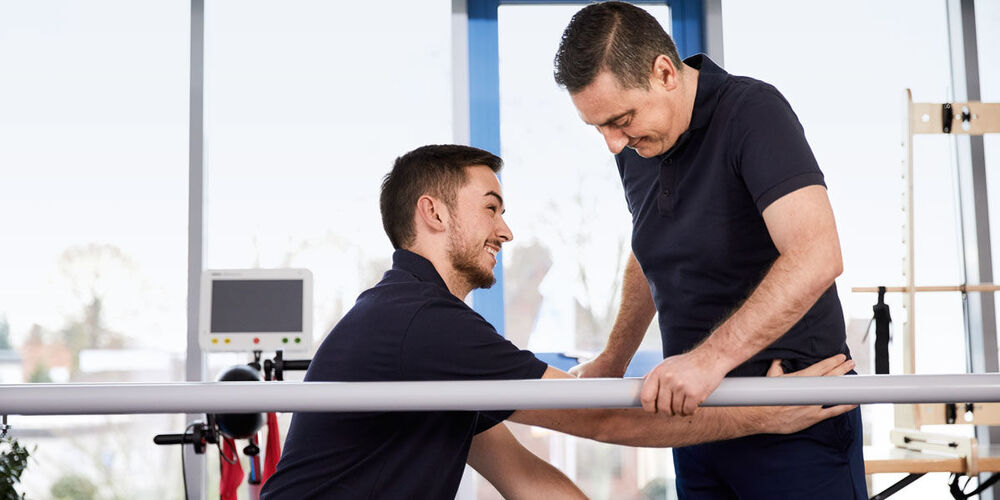Therapeut übt mit Patient das Gehen an der Stange.