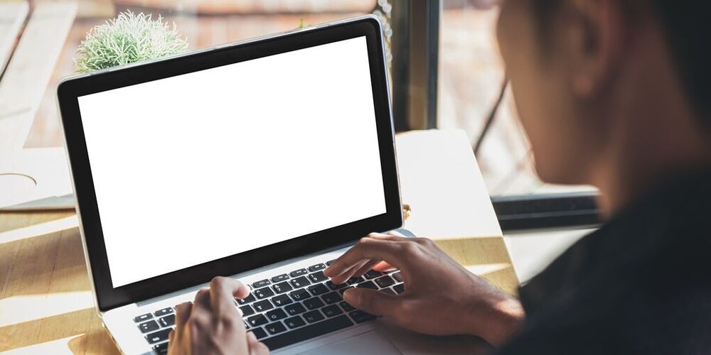 Ein Mann sitzt mit seinem Laptop vor einem Fenster, durch welches die Sonne scheint und tippt etwas auf diesem.