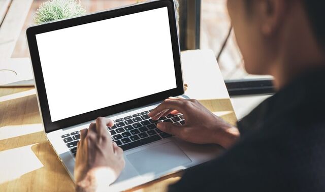 Ein Mann sitzt mit seinem Laptop vor einem Fenster, durch welches die Sonne scheint und tippt etwas auf diesem.