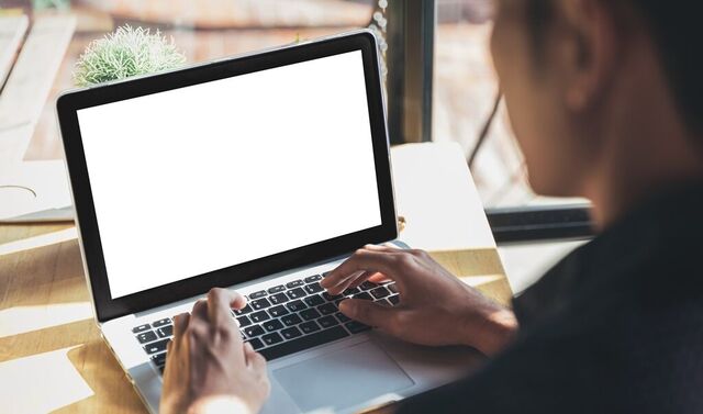Ein Mann sitzt mit seinem Laptop vor einem Fenster, durch welches die Sonne scheint und tippt etwas auf diesem.