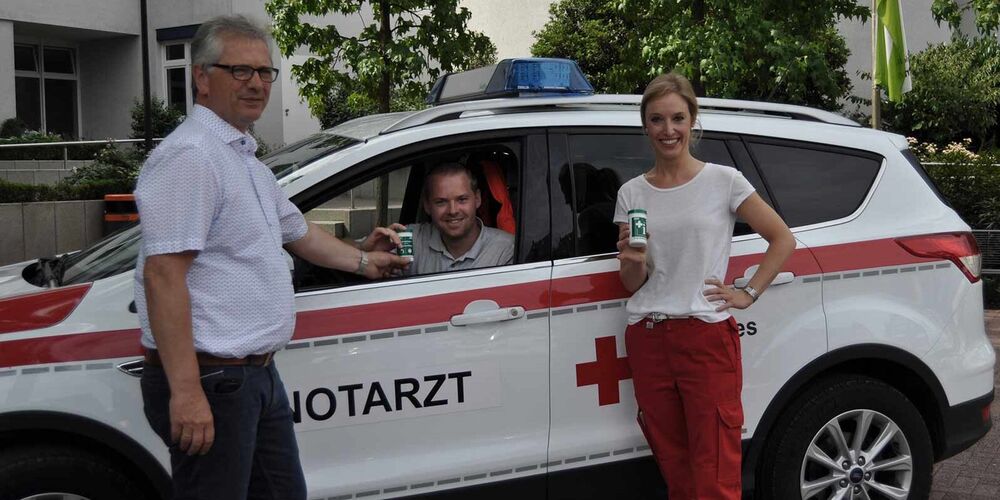 Team der Notärzte. Ein Mann sitzt in einem Auto auf dem Notarzt steht und vor diesem steht ein weiterer Mann und eine Frau. Alle drei schauen in die Kamera und lächeln. Außerdem halten sie zwei Notfalldosen in der Hand. 