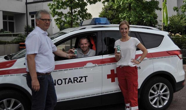 Team der Notärzte. Ein Mann sitzt in einem Auto auf dem Notarzt steht und vor diesem steht ein weiterer Mann und eine Frau. Alle drei schauen in die Kamera und lächeln. Außerdem halten sie zwei Notfalldosen in der Hand. 