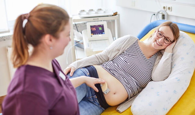Eine Schwangere liegt seitwärts auf einem Patientenbett. Eine Hebamme untersucht währenddessen den Herzschlag des Babys im Bauch.