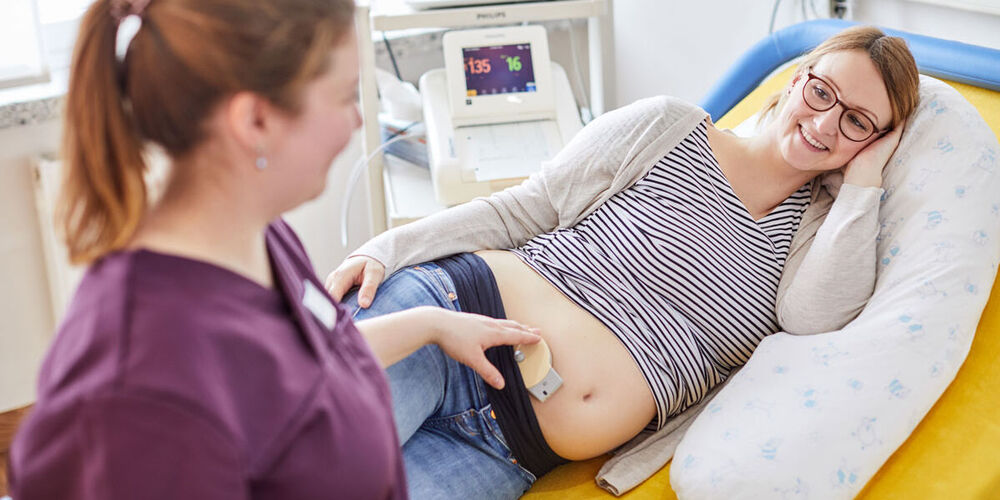 Eine Schwangere liegt seitwärts auf einem Patientenbett. Eine Hebamme untersucht währenddessen den Herzschlag des Babys im Bauch.