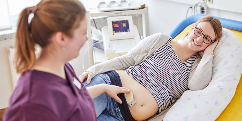 Eine Schwangere liegt seitwärts auf einem Patientenbett. Eine Hebamme untersucht währenddessen den Herzschlag des Babys im Bauch.
