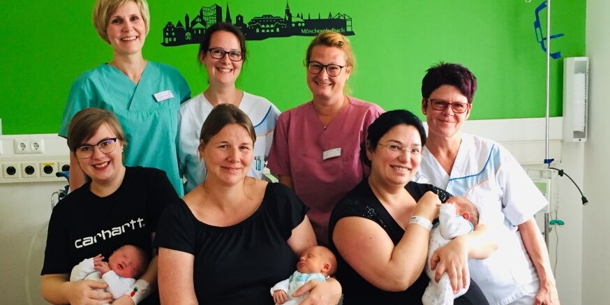 Geburtssaal der Klinik Niederrhein. Auf einem großen Bett sitzen drei Mütter mit ihren Neugeborenen. Hinter ihnen sitzen vier Geburtshelferinnen. 