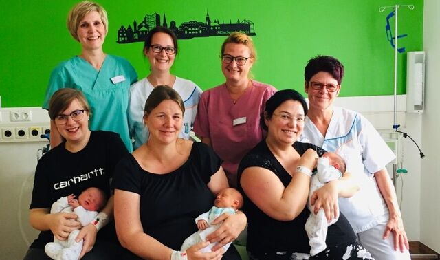Geburtssaal der Klinik Niederrhein. Auf einem großen Bett sitzen drei Mütter mit ihren Neugeborenen. Hinter ihnen sitzen vier Geburtshelferinnen. 
