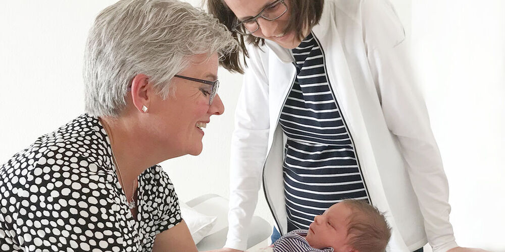 Eine Frau hält ein Neugeborenes in den Armen und stützt sich dabei auf einem Wickeltisch ab. Neben ihr steht eine Ärztin und schaut ebenfalls auf das Neugeborene.