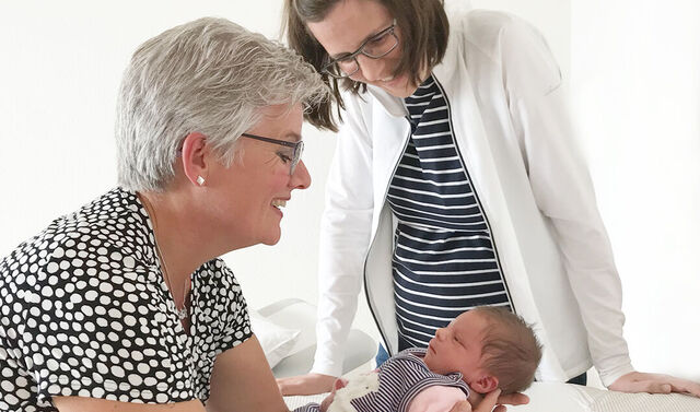 Eine Frau hält ein Neugeborenes in den Armen und stützt sich dabei auf einem Wickeltisch ab. Neben ihr steht eine Ärztin und schaut ebenfalls auf das Neugeborene.