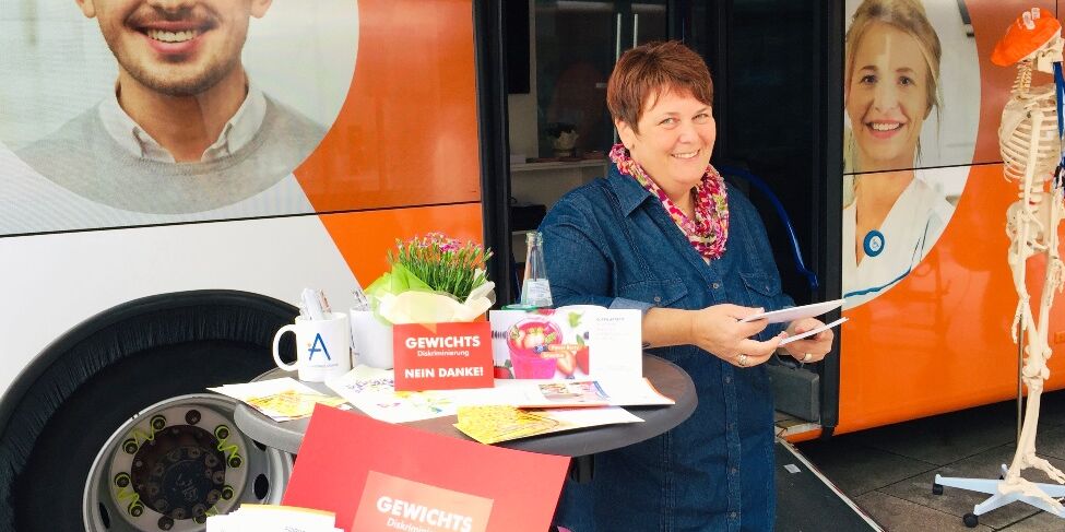 Eine Frau steht vor einem Bus mit Werbematerialien und Flyern. Auf den Flyern steht Gewichtsdiskriminierung nein danke! 