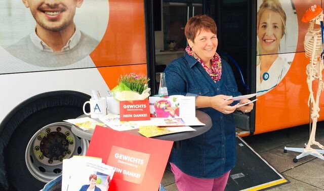 Eine Frau steht vor einem Bus mit Werbematerialien und Flyern. Auf den Flyern steht Gewichtsdiskriminierung nein danke!