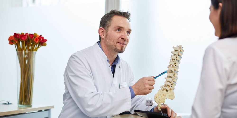 Ein Arzt sitzt einer Patientin gegenüber und befindet sich mit dieser im Gespräch. Hierbei zeigt er auf das Modell einer Wirbelsäule. 