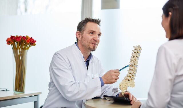 Ein Arzt sitzt einer Patientin gegenüber und befindet sich mit dieser im Gespräch. Hierbei zeigt er auf das Modell einer Wirbelsäule. 
