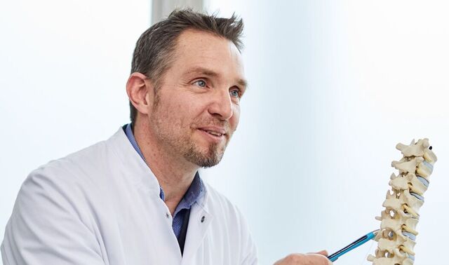 Ein Arzt sitzt einer Patientin gegenüber und befindet sich mit dieser im Gespräch. Hierbei zeigt er auf das Modell einer Wirbelsäule. 