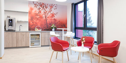 Moderne Sitzlounge mit drei Loungestühlen sowie einem Tisch. Außerdem stehen im Hintergrund ein Stehtisch mit drei Barstühlen. Außerdem sind im Hintergrund eine Kaffeemaschine sowie ein Waschbecken und ein Kühlschrank. 