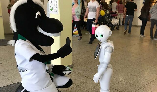 Ein Maskottchen in Form eines Pferdes kniet vor einem Roboter und hält eine Hand zum einschlagen hoch.