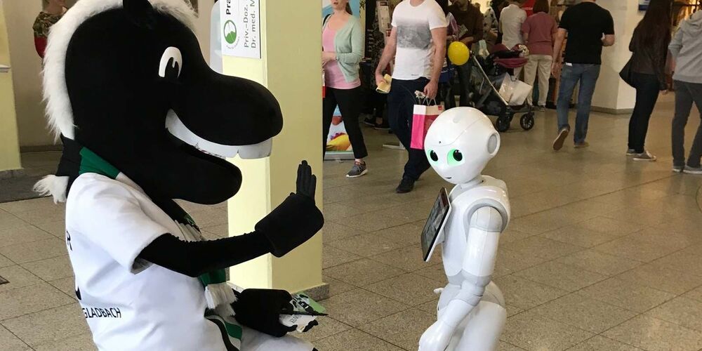 Ein Maskottchen in Form eines Pferdes kniet vor einem Roboter und hält eine Hand zum einschlagen hoch.