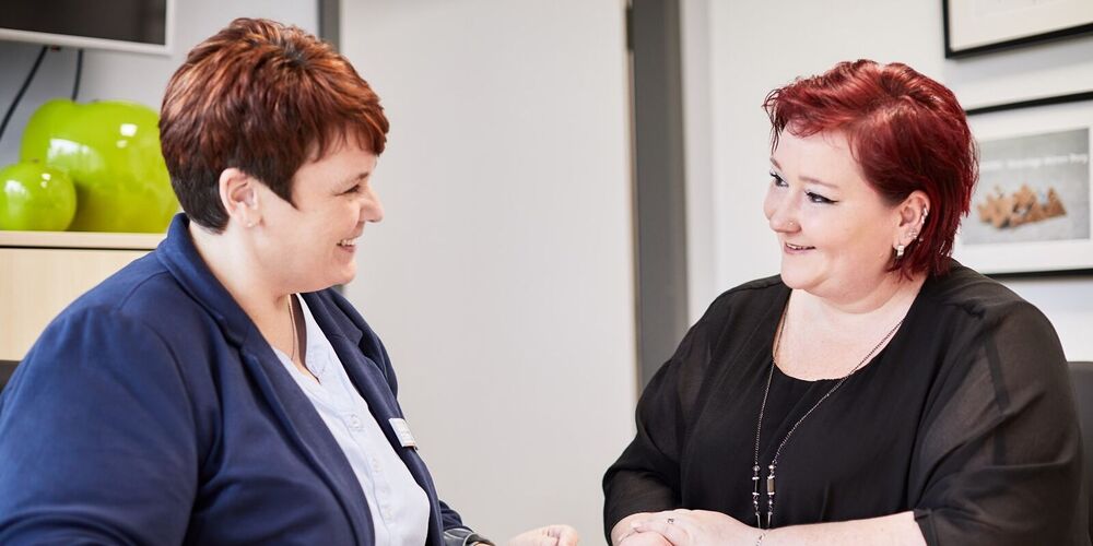 Zwei Frauen sitzen an einem Tisch und unterhalten sich. Eine Frau hält einen Stift in der Hand und zeigt etwas auf einem Zettel. Es handelt sich scheinbar um eine Beratungssituation.