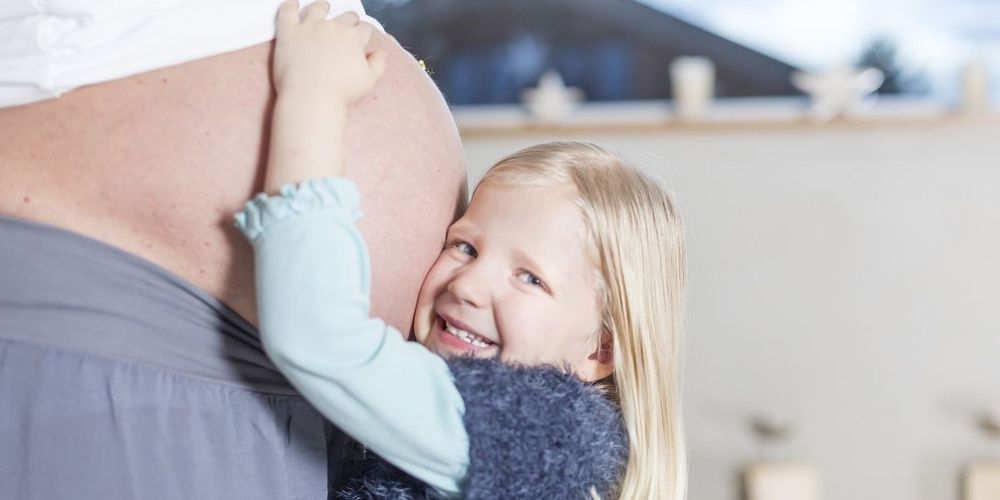 Ein kleines Mädchen steht vor dem Babybauch ihrer Mutter und hält dieses umarmt und drückt ihre Wange an diesen. Dabei schaut sie lächelnd in die Kamera.
