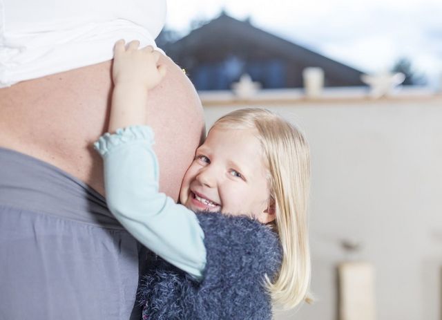 Ein kleines Mädchen steht vor dem Babybauch ihrer Mutter und hält dieses umarmt und drückt ihre Wange an diesen. Dabei schaut sie lächelnd in die Kamera.