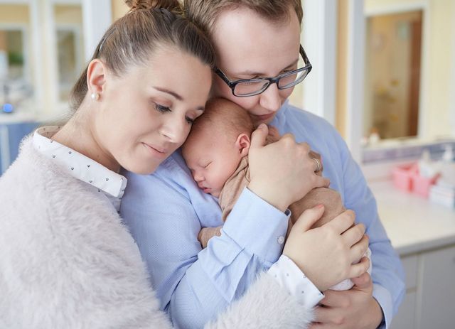 Ein junges Paar das sich umarmt und ihr neugeborenes Baby auf dem Arm hält.
