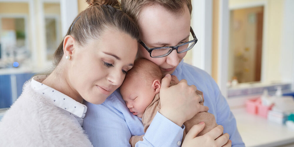 Ein junges Paar das sich umarmt und ihr neugeborenes Baby auf dem Arm hält.