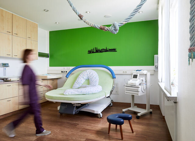 Kreissaal mit einem Geburtsbett. Eine Geburtshilferin läuft durch das Zimmer.