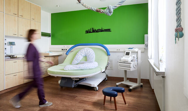 Kreissaal mit einem Geburtsbett. Eine Geburtshilferin läuft durch das Zimmer. 
